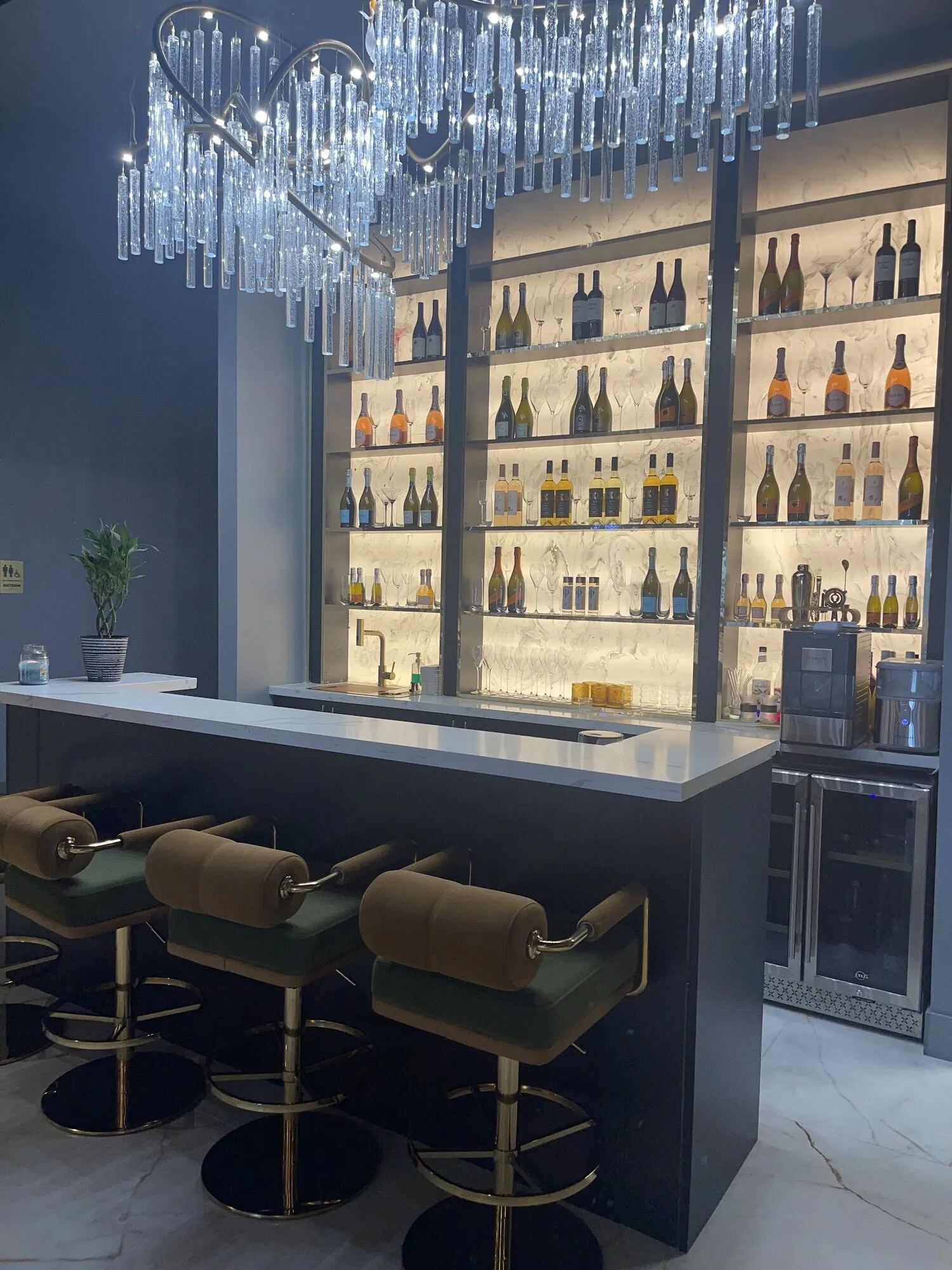 The bar at Zara Nail Bar Morrison: backlit shelves of bottles under a crystal chandelier, with green leather stools at the counter.